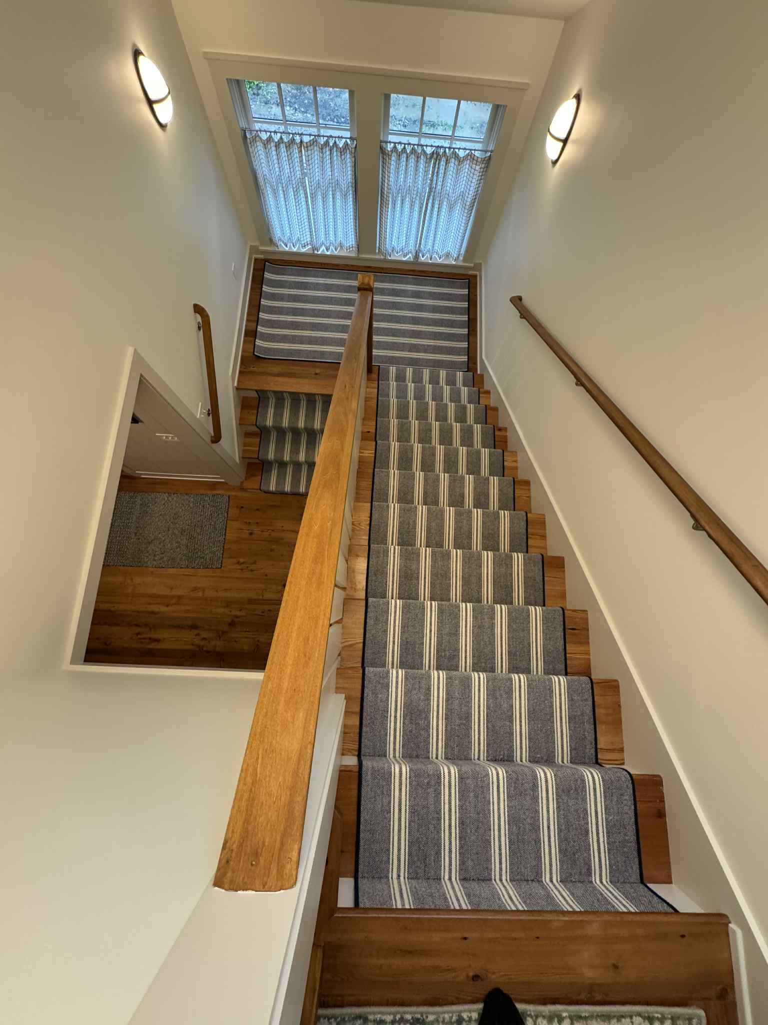 Carpeted stair runner with gray and tan stripes installed over wooden stair treads in a residential stairwell with wooden handrails.