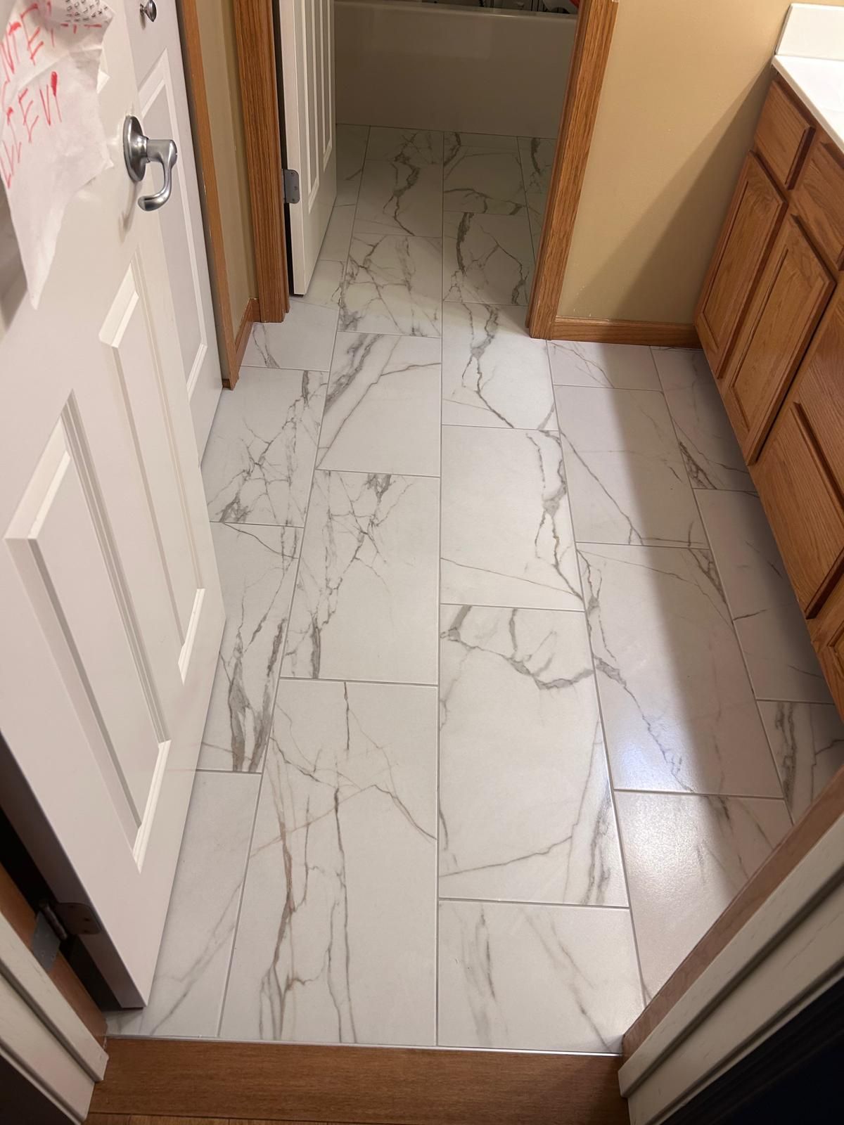 Light gray and white marble-look tile flooring in plank pattern installed in a bathroom with wood-frame vanity and white walls.