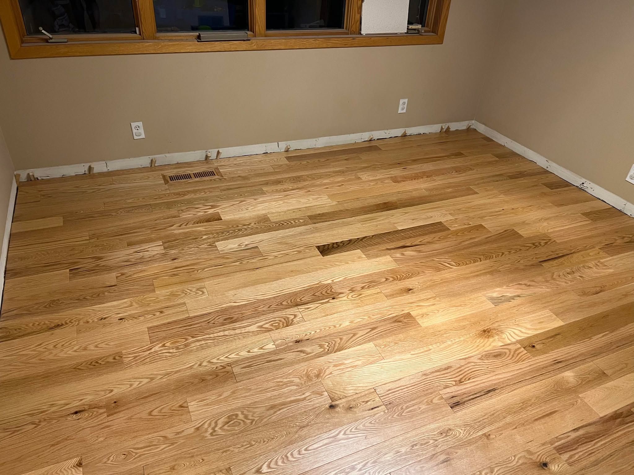 Newly installed light tan hardwood plank flooring in a bedroom with white trim and heat registers visible.