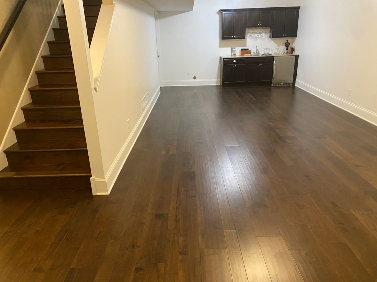 Basement with dark hardwood plank flooring, stairs on left, and small kitchenette with dark cabinets.