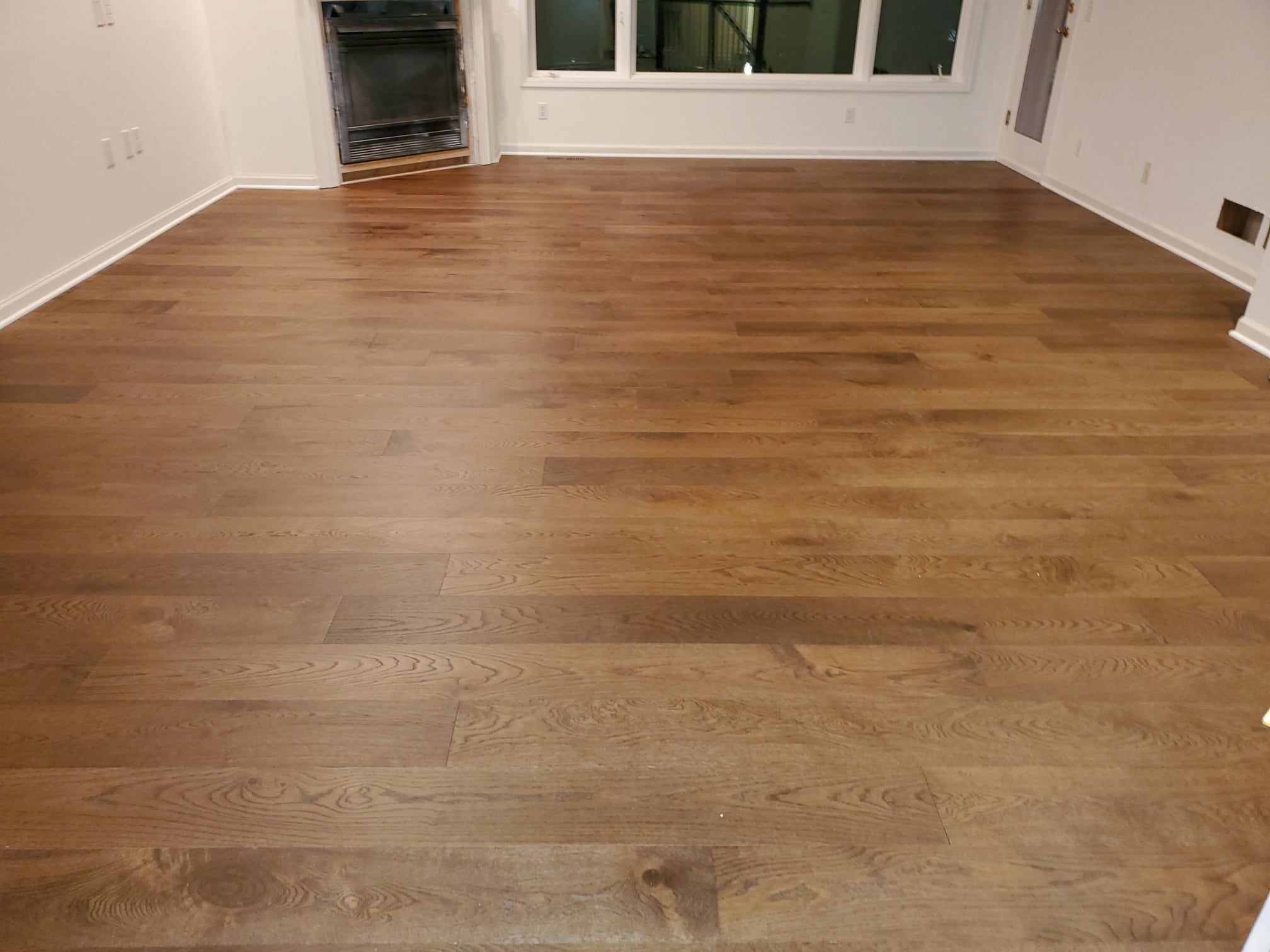 Medium brown hardwood plank flooring in an empty living room with white walls and large windows.