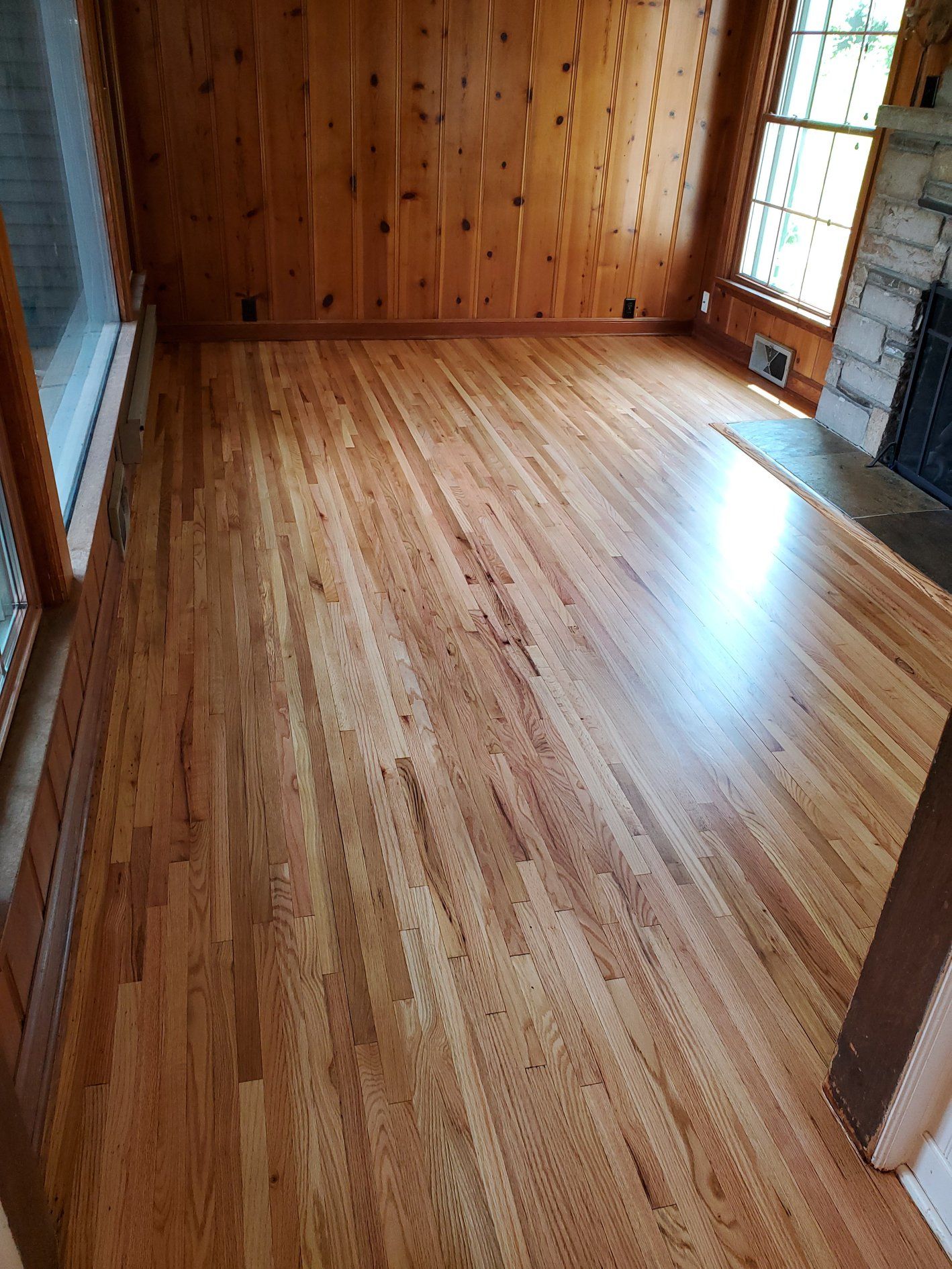 Newly refinished red oak hardwood flooring in plank pattern with medium tan-brown finish in a bedroom with knotty pine ceiling.