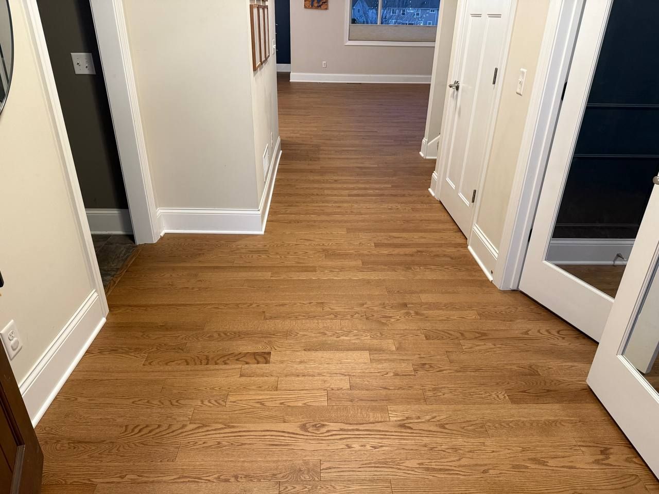 Medium-toned oak hardwood plank flooring installed in a hallway with white trim and multiple doorways.