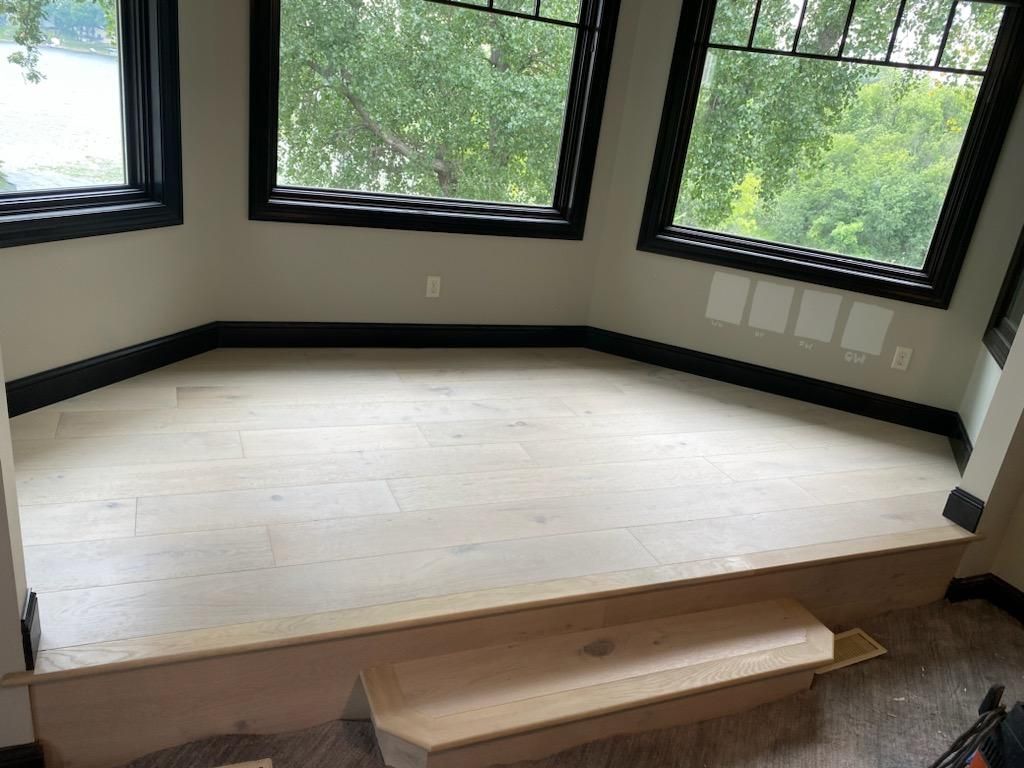 Light-colored hardwood plank flooring installed on an elevated platform in a sunlit bedroom with black trim and windows.