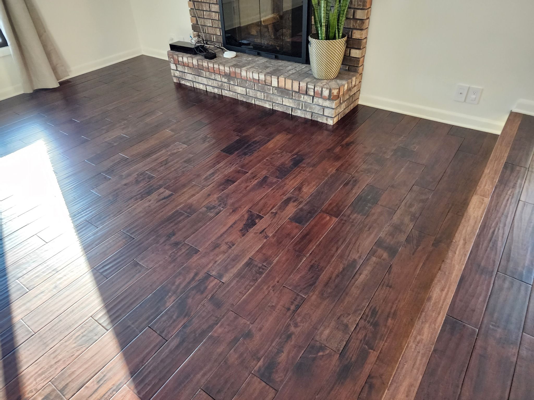 Dark hardwood plank flooring in living room with brick fireplace surround and bright sunlight reflection.