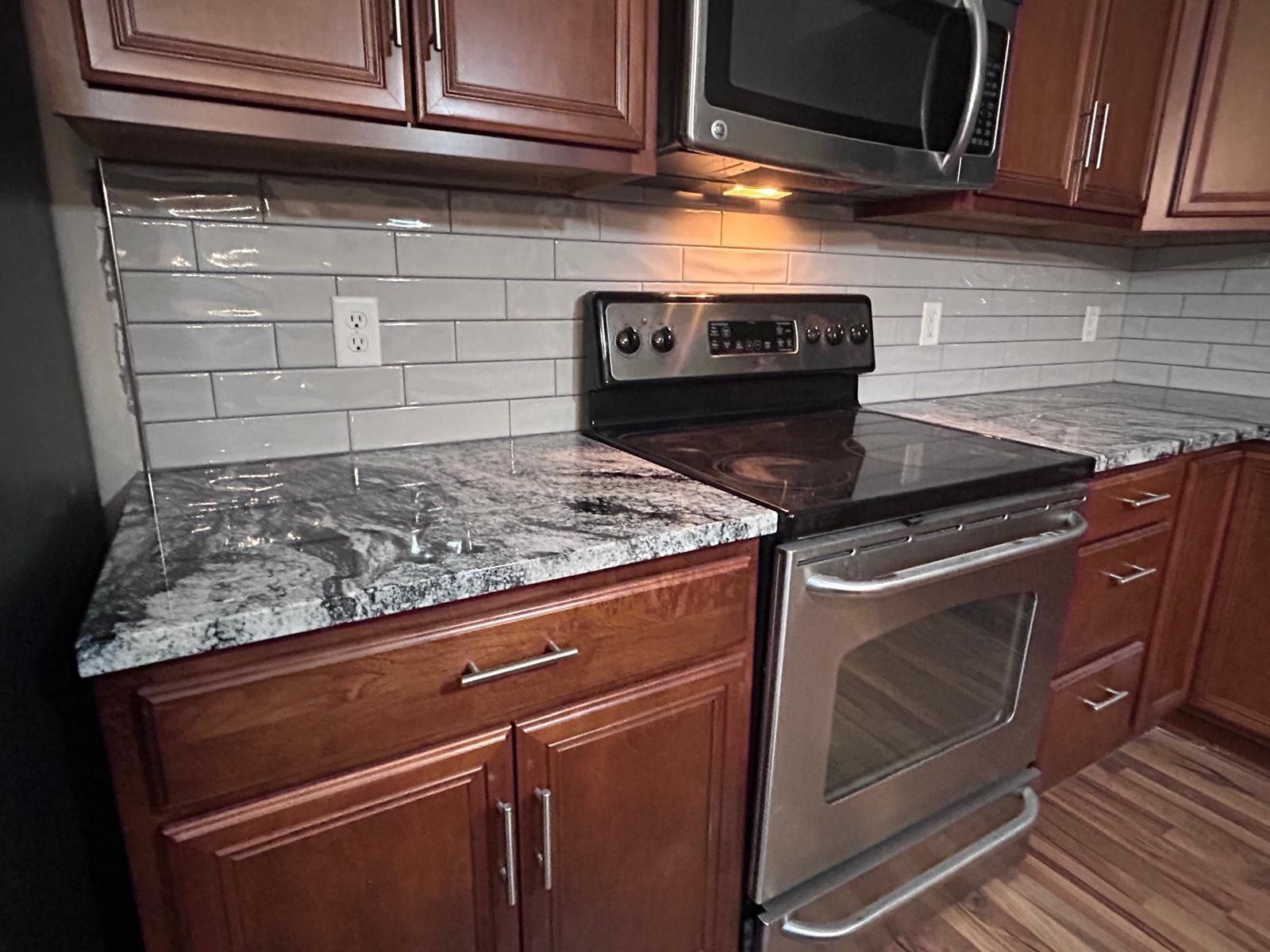 White subway tile backsplash with grout lines installed above a stove in a kitchen with cherry cabinets and granite countertop.