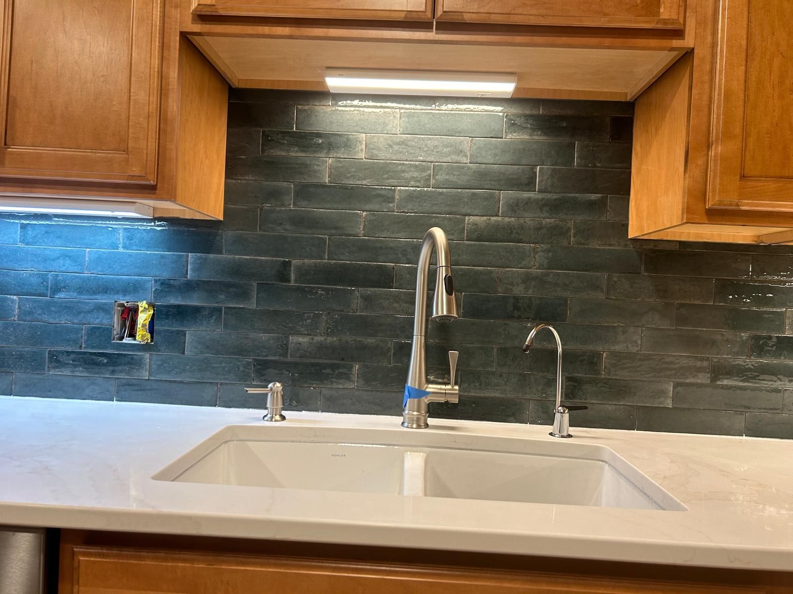 Dark subway tile backsplash above kitchen sink with stainless steel faucet and cream countertop.