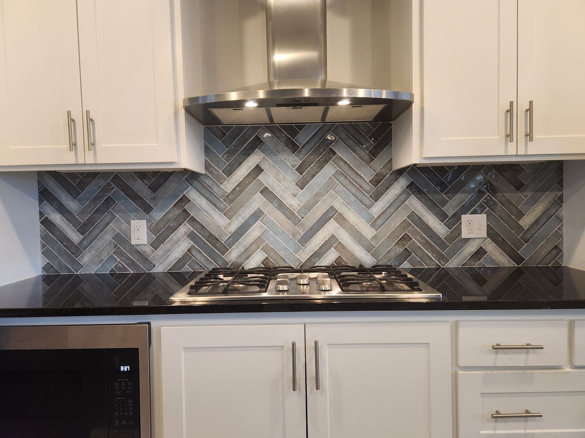 Kitchen backsplash with gray and white herringbone tile pattern above a gas cooktop.