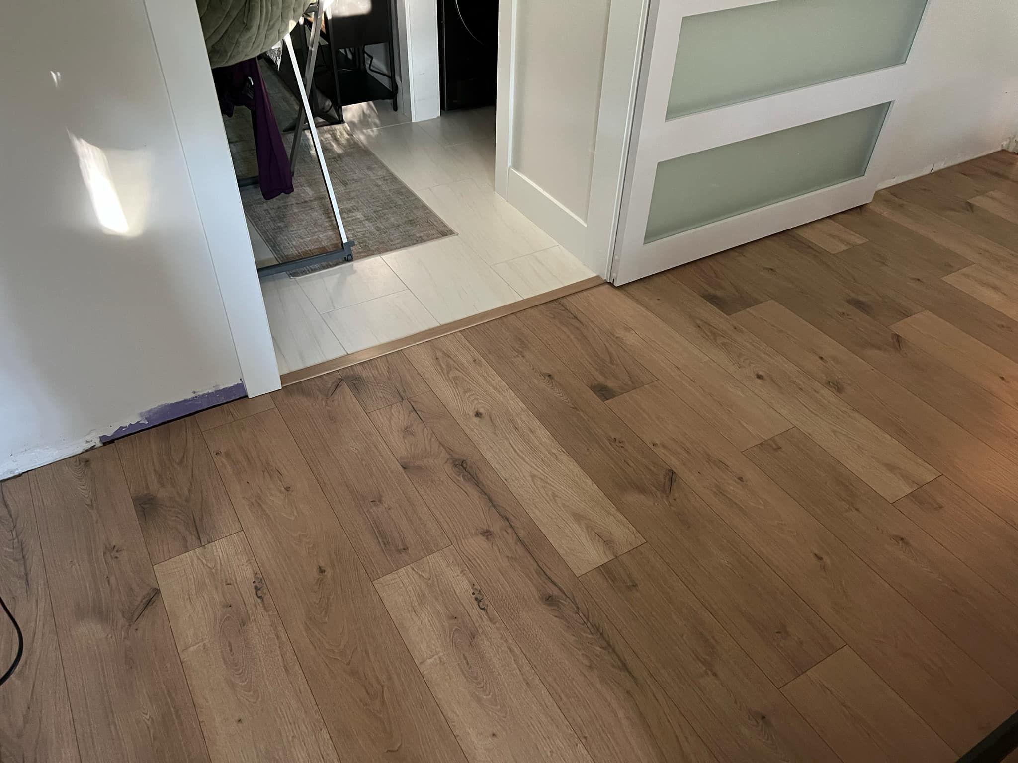 Hallway with medium-toned laminate plank flooring transitioning to a light tile entry area.