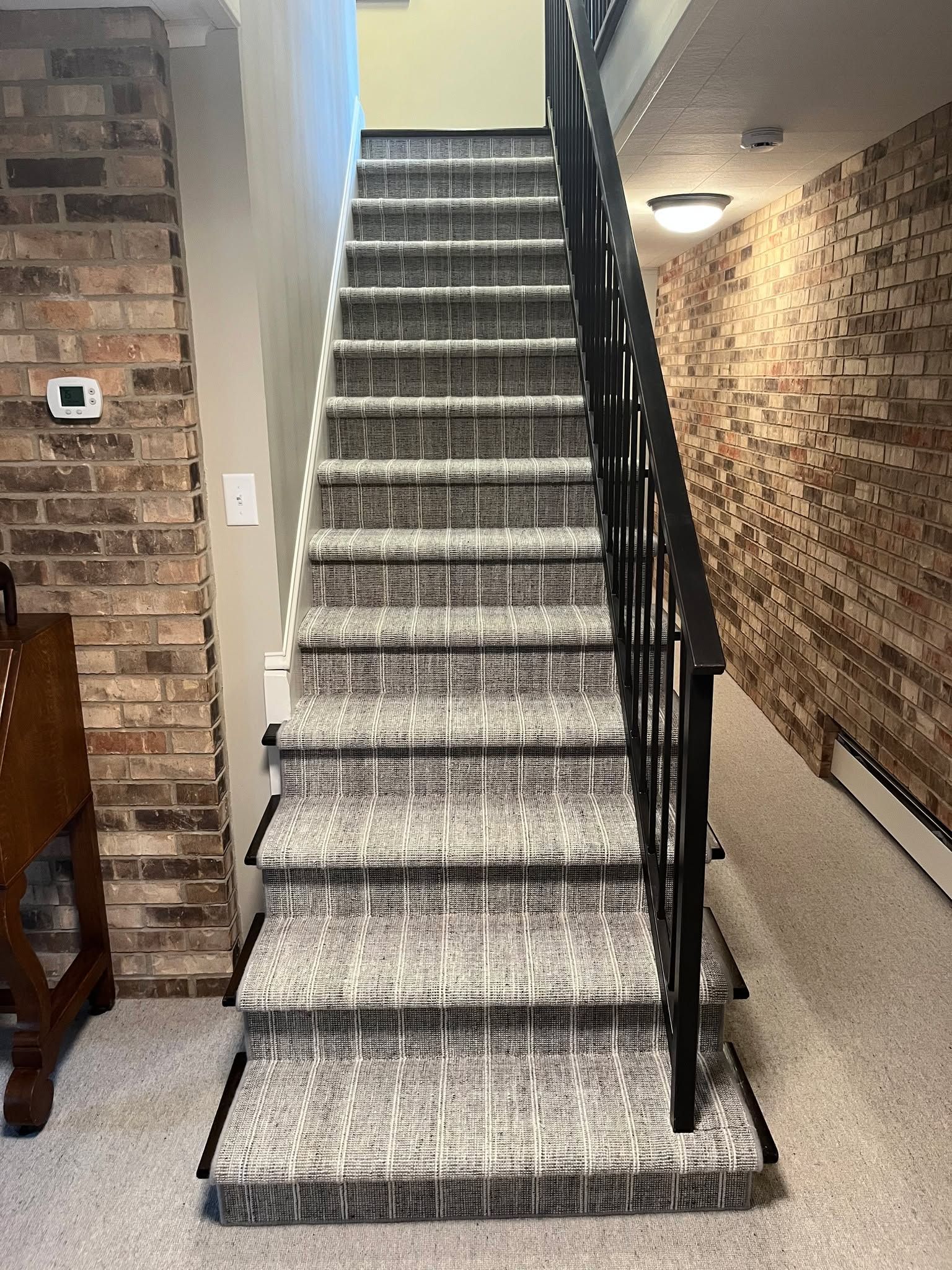 Carpeted staircase with gray and white patterned runner and black metal handrail in basement with brick walls.