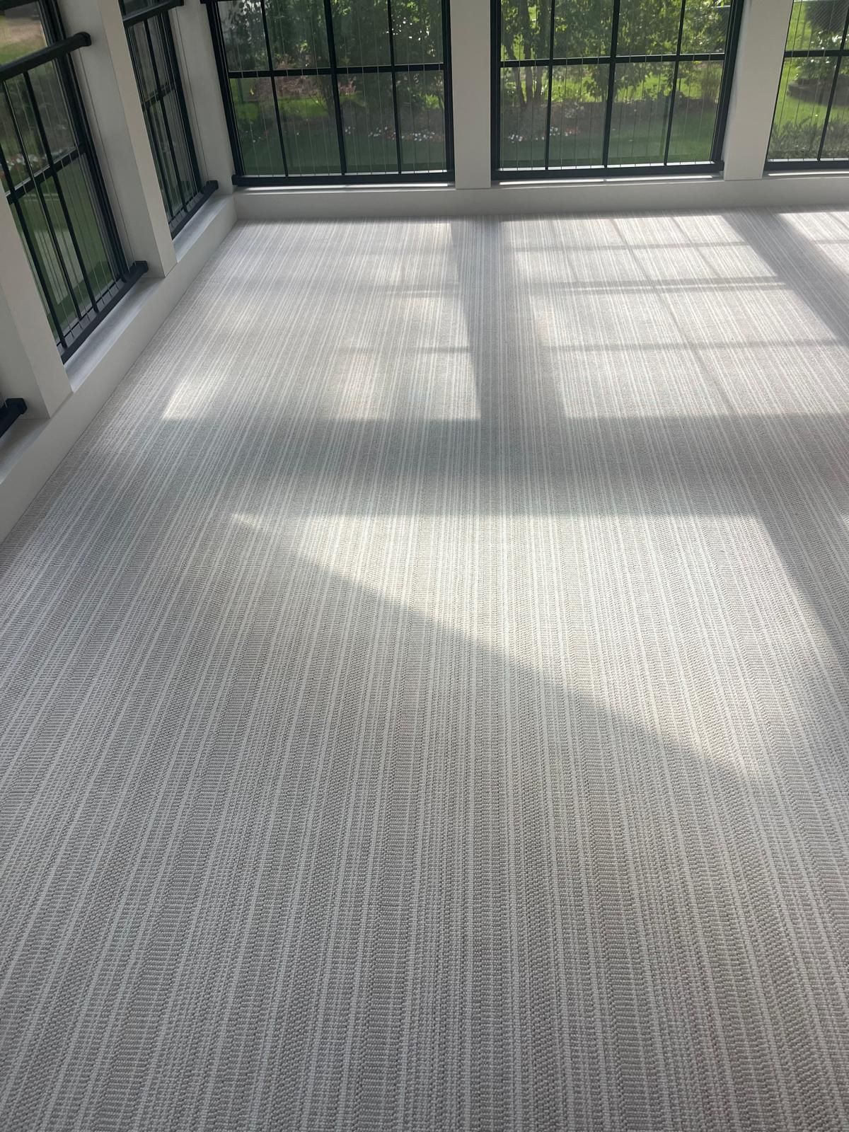 Light gray indoor-outdoor carpet with linear texture installed in a sunlit room with large windows.
