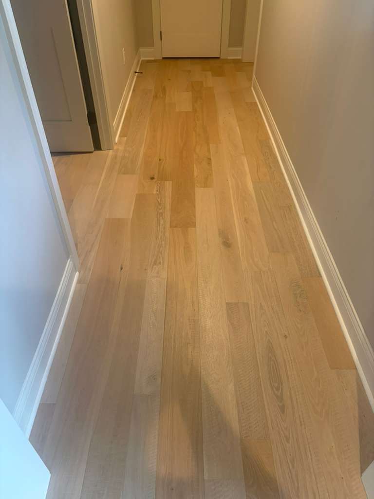 Light-toned hardwood plank flooring installed in a narrow hallway with white baseboards and interior doors.